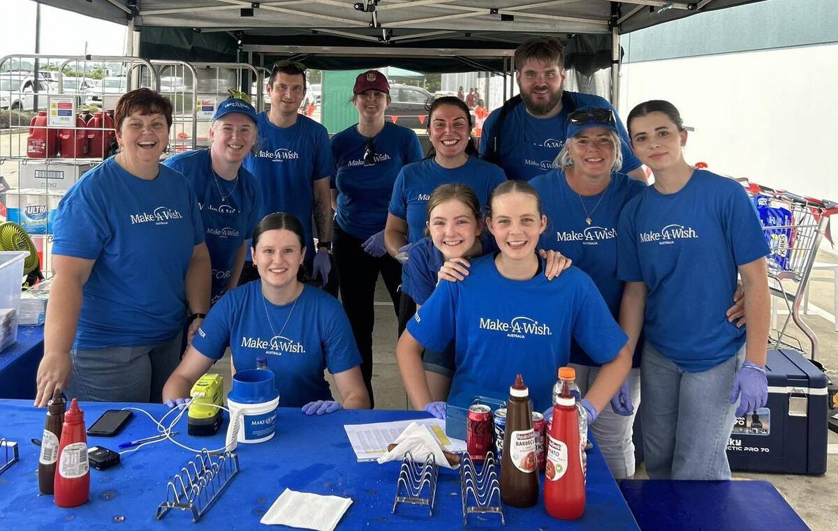 Eleven Make-A-Wish volunteers pose together under a tent in the parking lot of a Bunnings Warehouse. They are all wearing blue Make-A-Wish t-shirts and smiling at the camera. Condiments sit on the table in front of them, ready to be used in the day's sausage sizzle fundraiser.