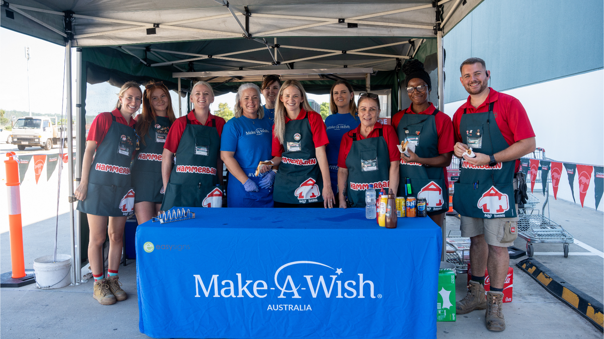 Ten Make-A-Wish volunteers standing together behind a table with a blue Make-A-Wish tablecloth. The volunteers are wearing 'Hammerbarn' aprons and smiling at the camera. They are posed under a tent in a Bunnings parking lot, where the BBQ is hosted.