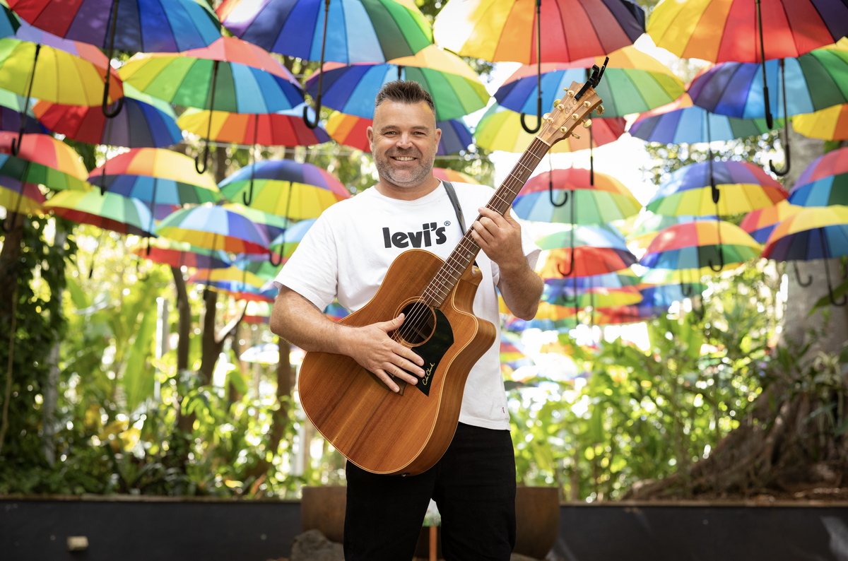 Man with guitar stands in front of rainbow umbrellas