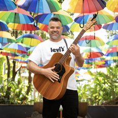 Man with guitar stands in front of rainbow umbrellas