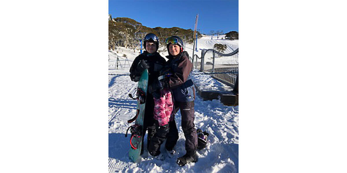 Harry holding a snowboard standing on a snowy mountain with a ski field in the background