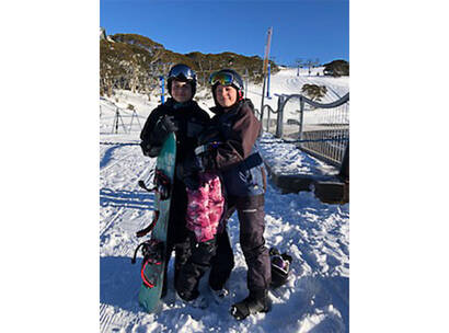Harry holding a snowboard standing on a snowy mountain with a ski field in the background