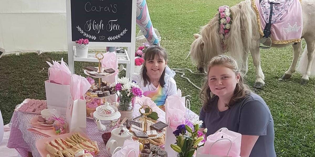 Make-A-Wish Australia wish child Cara sat with a friend at a table with a unicorn behind