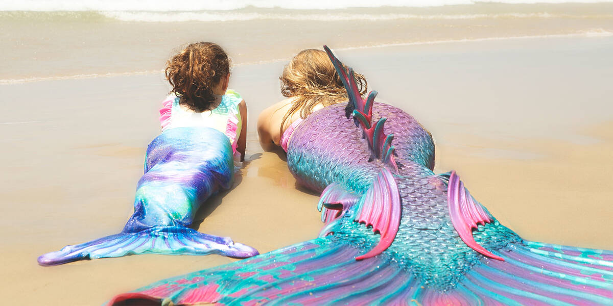 Make-A-Wish kid Jazmyn with a mermaid lie facing away from the camera so that you can see their colourful tails on the sand