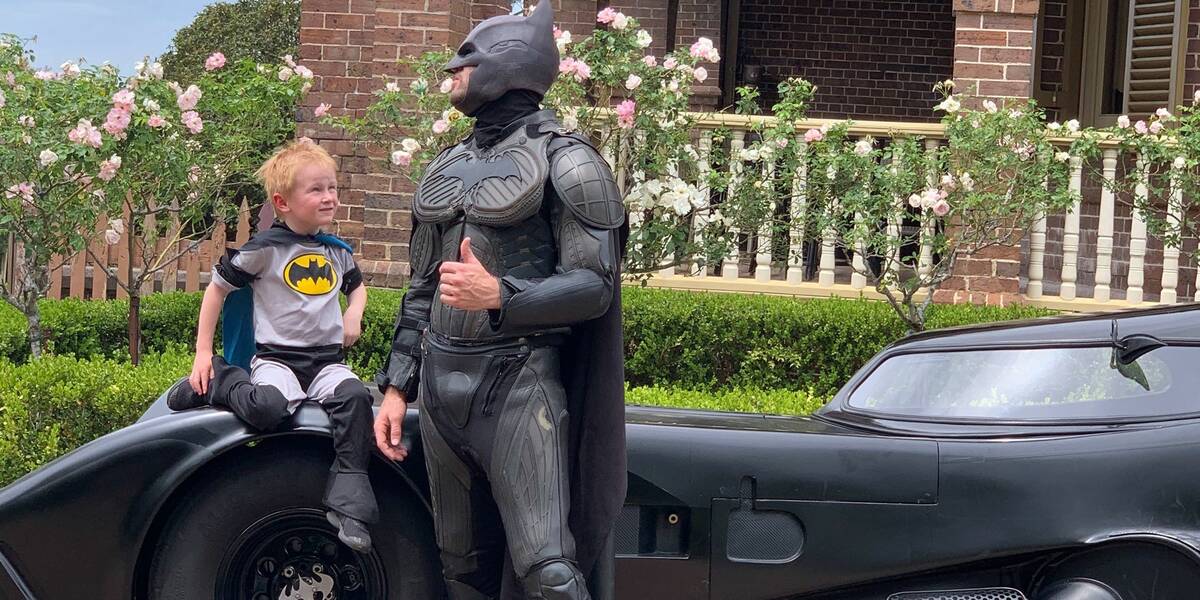 Batmas carries Make-A-Wish kid Rory and the two of them laugh while standing in front of the batmobile